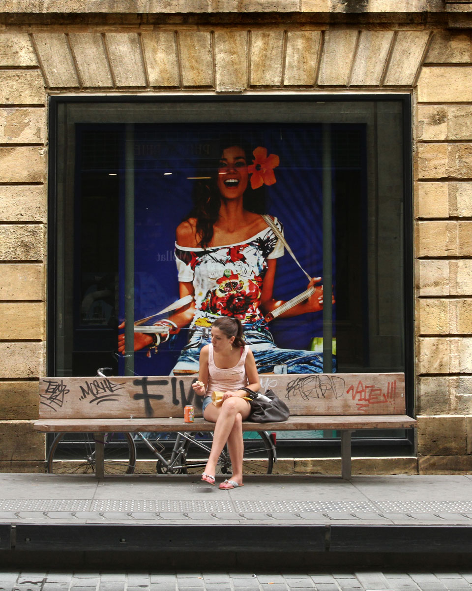 Eine Frau sitzt vor einem Schaufenster in Bordeaux, in dem ein Poster mit einem übergroßen Frauenportrait, das über die sitzende Frau hinweg sieht und lacht, als gehörten beide zusammen.