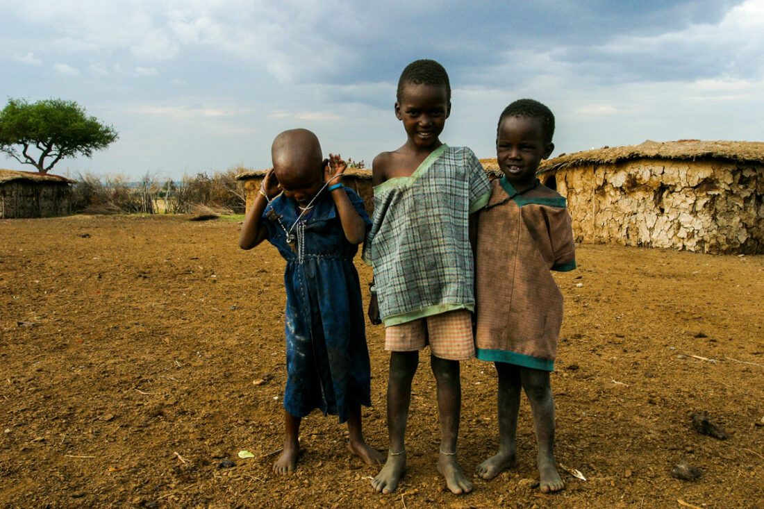 drei kleine Masai Kinder stehen stolz und posieren vor den Hütten eines Runddorfes.