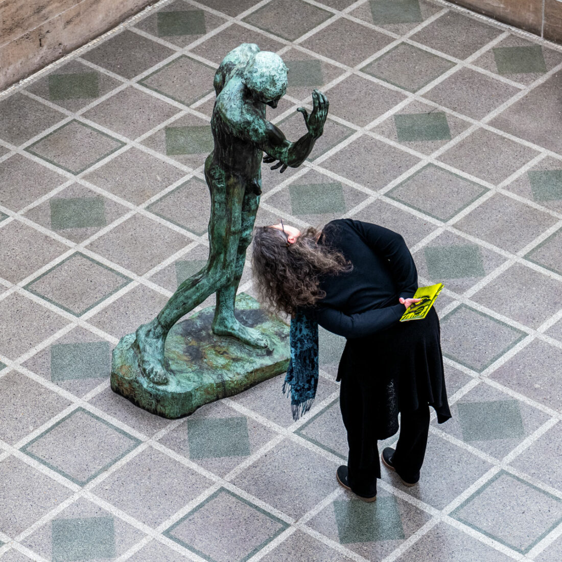 Eine Ausstellungsbesucherin versucht einer Statue ins nach unten gesenkte Gesicht zu schauen und muss sich dafür nach unten beugen, um so in das Gesicht der Statue blicken zu können.