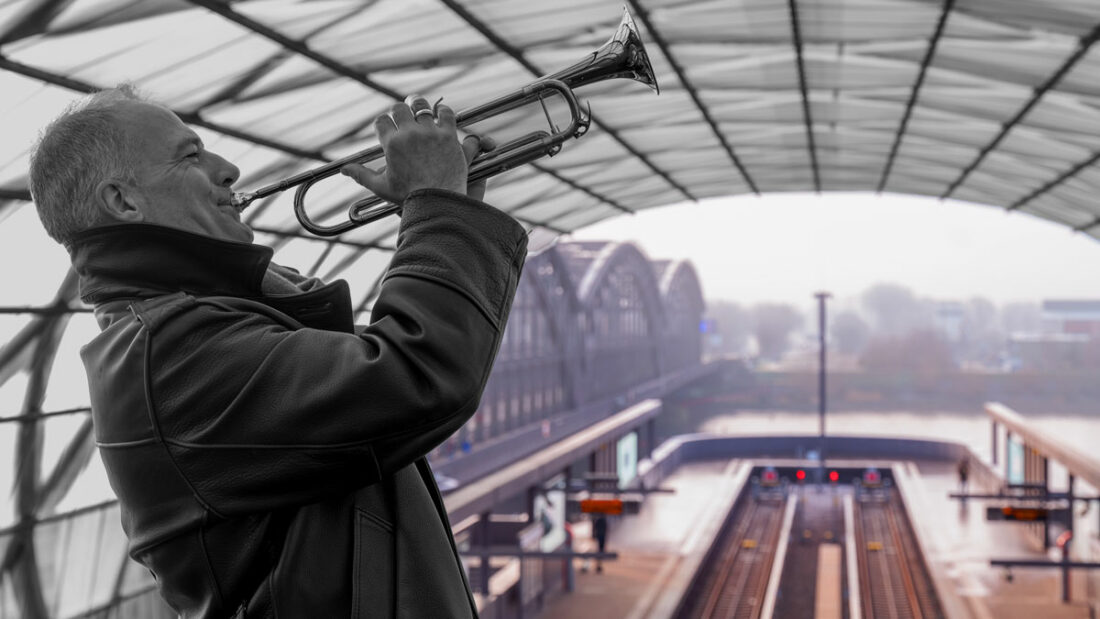ein Trompeter im U-Bahnhof Elbbrücken. Im Hintergrund die Elbbrücken. Der Trompeter und ds Bahnhofsdach sind schwarz-weiß, der Hintergrund in Farbe.