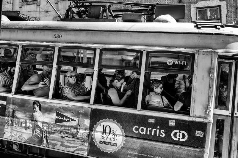 Schwarz-Weiss-Aufnahme einer vorbeifahrenden Strassenbahn in Lissabon mit herausschauenden Fahrgästen, die direkt in die Kamera blicken