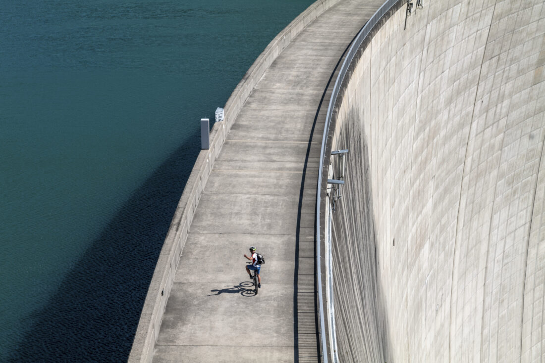 Ein Radfahrer auf der Staumauer des Stausees, wie er gerade zurückblickt. Neben ihm sein Schatten.